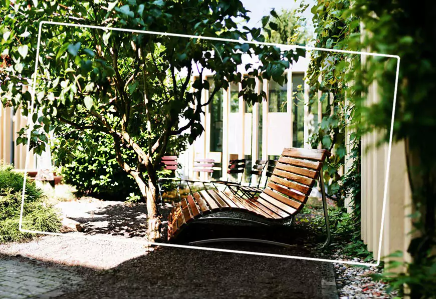 Nachhaltiges Tagungshaus Ein mit Baum, Büschen und Rankpflanzen begrünter Innenhof. eine Sitzecke mit Stühlen und eine Holz-Liege.