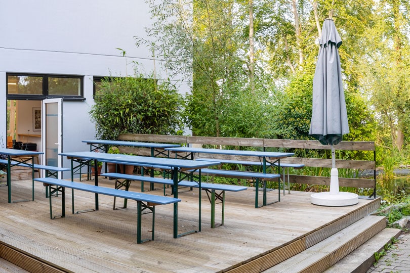 Terrasse der Jugendakademie mit Holztischen und Bänken sowie einem großem Sonnenschirm im Grünen.